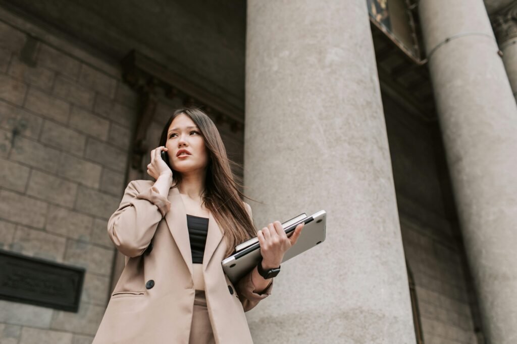 Asian businesswoman holding laptop and talking on mobile phone outside a modern building.